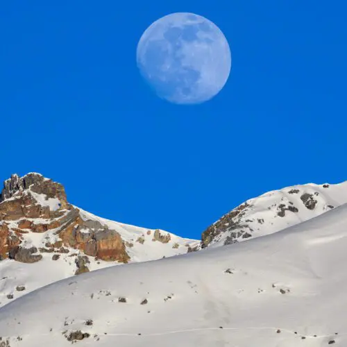 Grosser runder Mond über einem Schneerücken, durch den ein Wildwechsel mit einer Reihe winziger Gämsen zieht