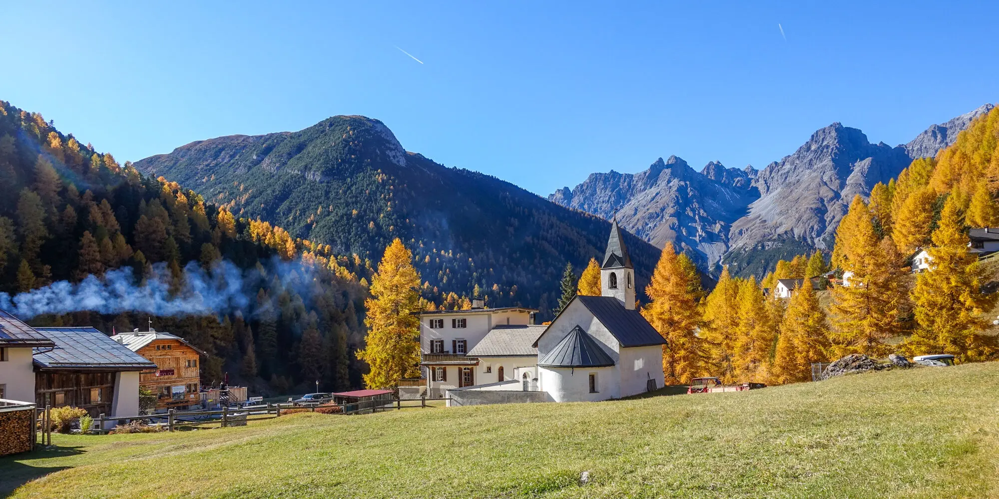 Herbstlandschaft im Ort S-charl, mit Kirche im Vordergrund und gelben Lärchen daneben
